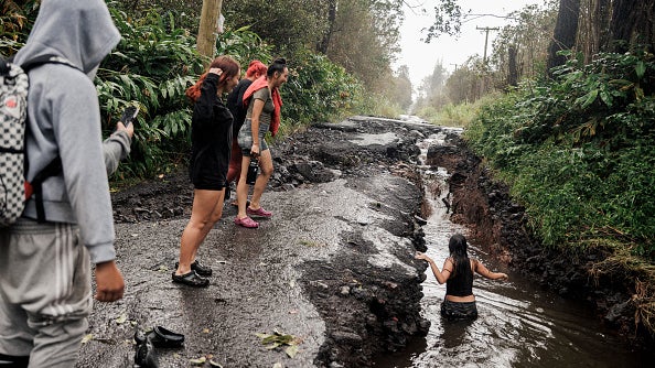 People stand on a damaged, flooded road while a child wades through water in a large crack in the pavement.