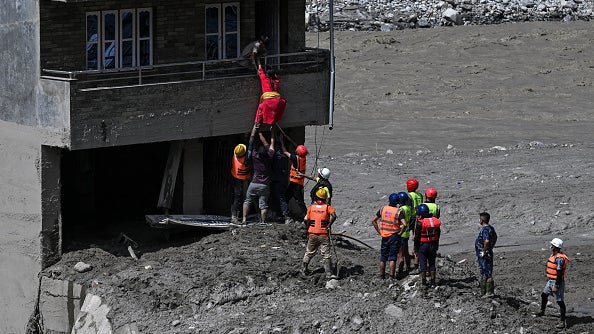 Rescue workers lift a person in a red suit toward a damaged building surrounded by flood debris and mud.