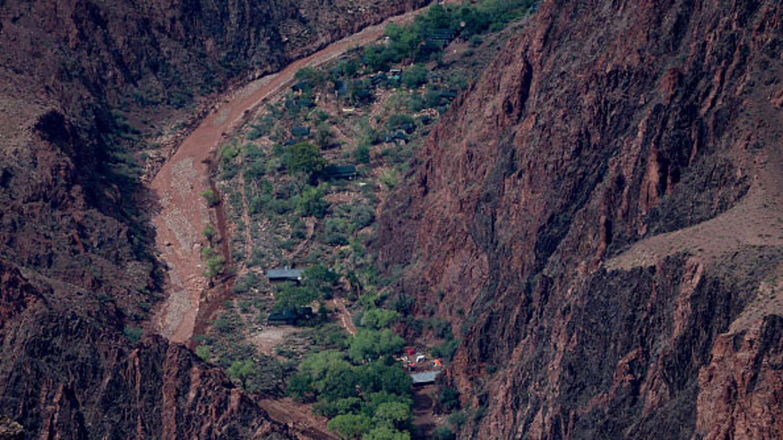 Aerial view of a muddy river winding through a canyon with green vegetation and small buildings nestled between steep rocky cliffs.