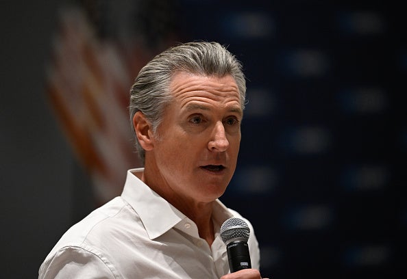 El gobernador Newsom habla por un micrófono en un evento, vestido con una camisa de cuello blanco.