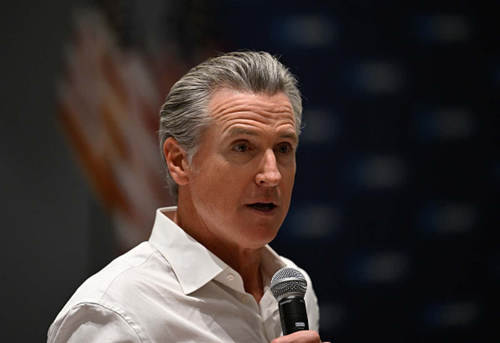El gobernador Newsom habla por un micrófono en un evento, vestido con una camisa de cuello blanco.