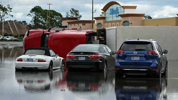 Flooded parking lot with an overturned red truck beside parked cars in front of a Publix store