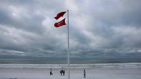 Two red warning flags on a pole flutter over a beach under a cloudy sky with people walking near the shore