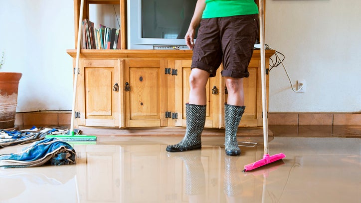 Person in rain boots standing on flooded floor beside wooden TV cabinet with mops and towels nearby.