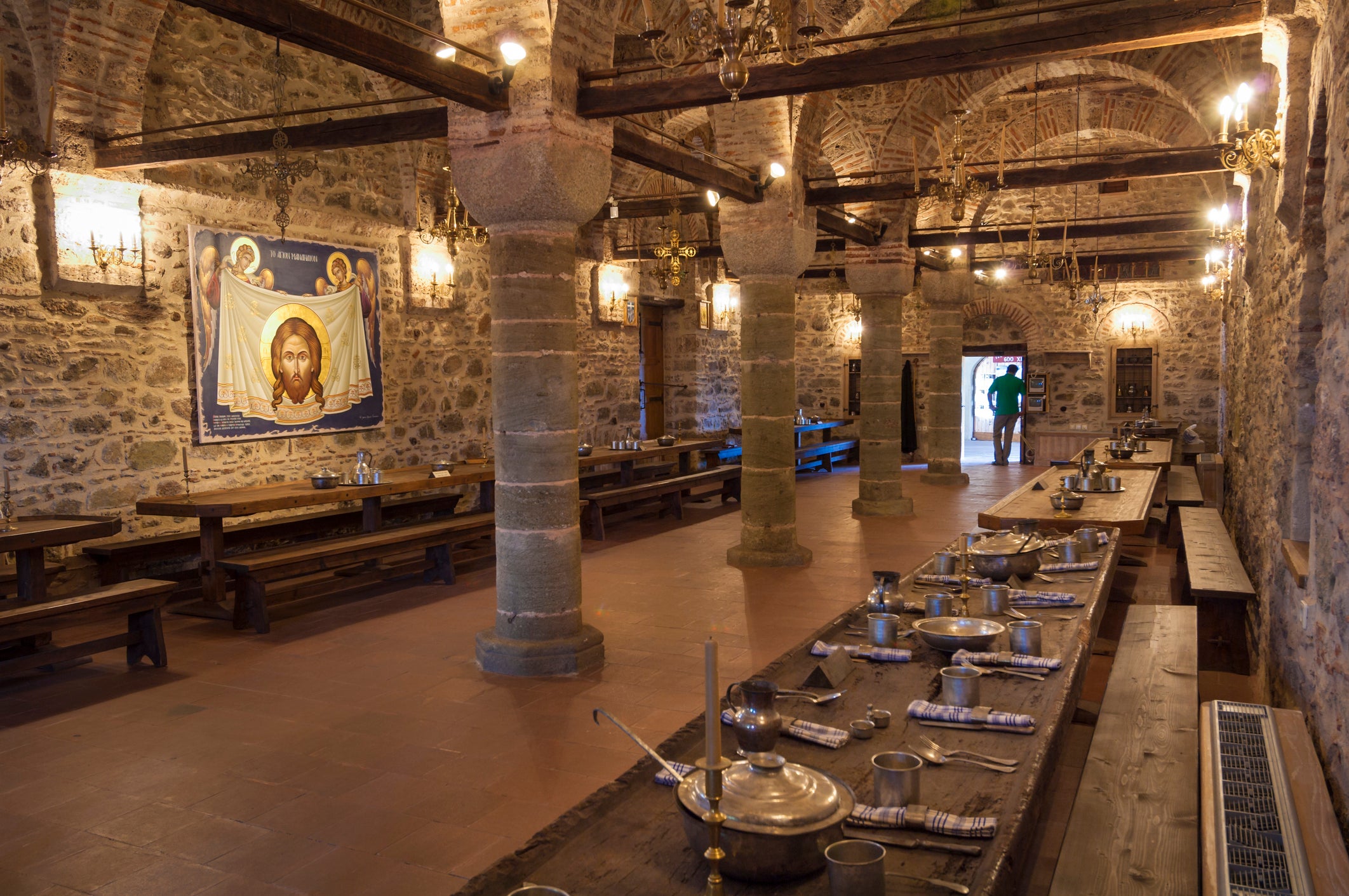 Monastery refectory with stone columns, vaulted brick ceiling, long wooden tables set with metal dishware, and a religious icon on the wall.