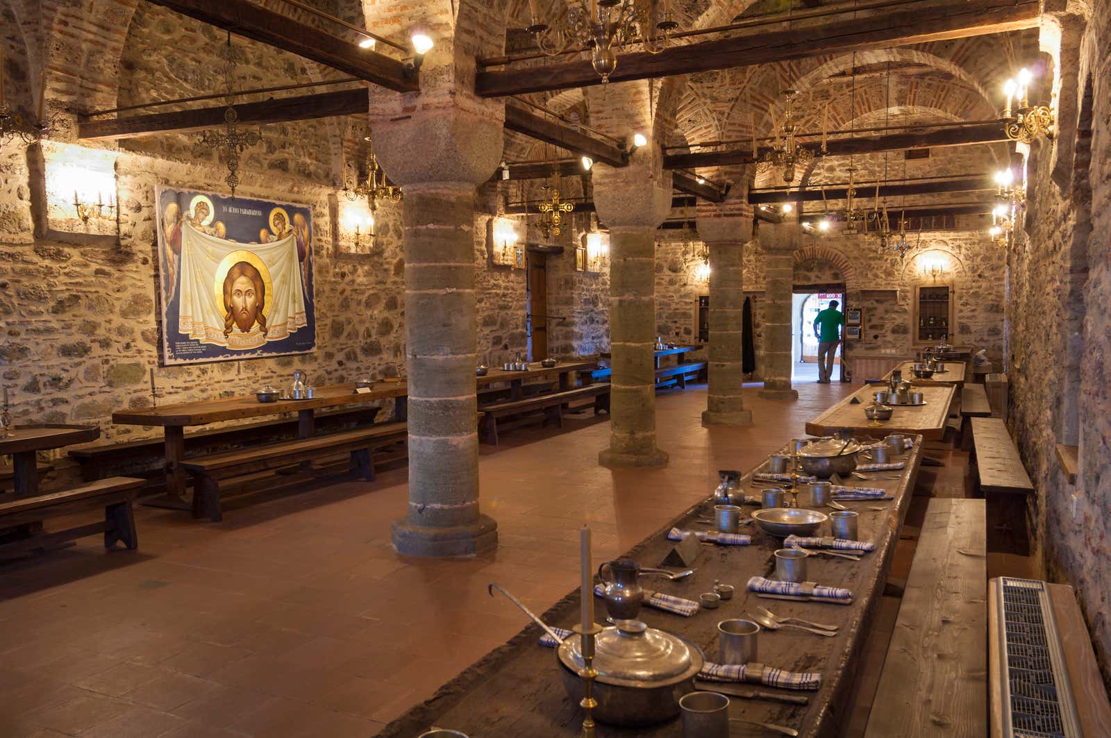 Monastery refectory with stone columns, vaulted brick ceiling, long wooden tables set with metal dishware, and a religious icon on the wall.