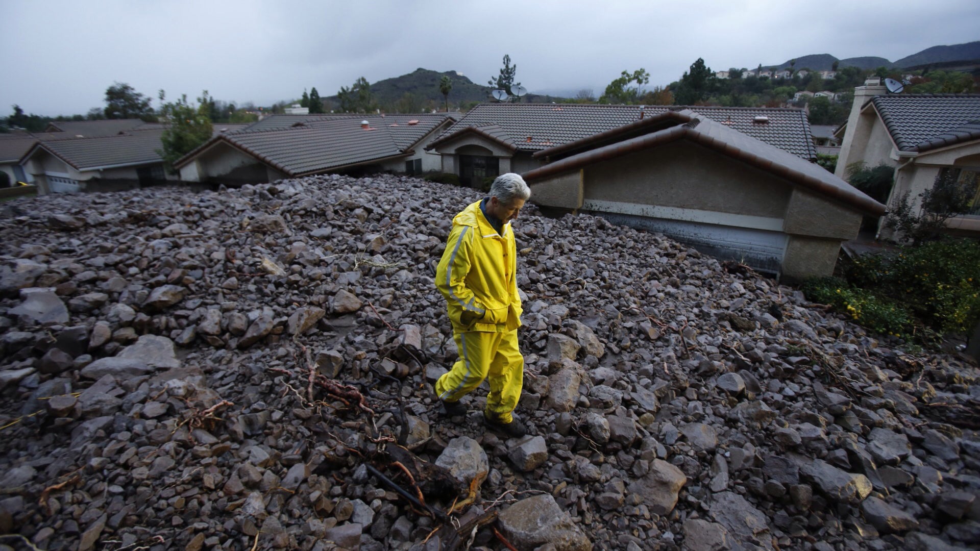 Person in yellow rain gear walking through mudslide debris field with damaged houses and mountains in background