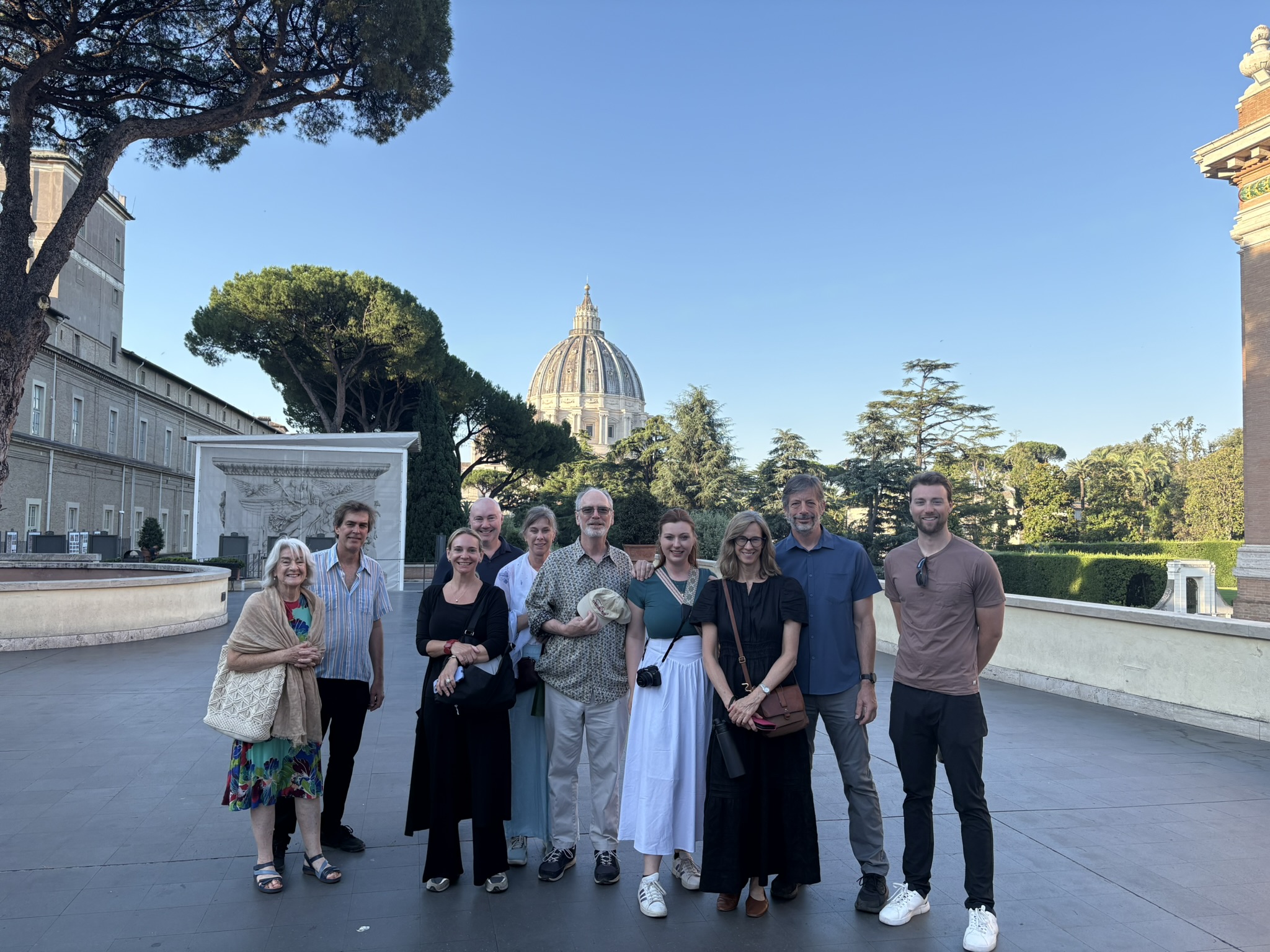 Nine people posing outdoors with St. Peter&rsquo;s Basilica dome in the background and trees framing the scene