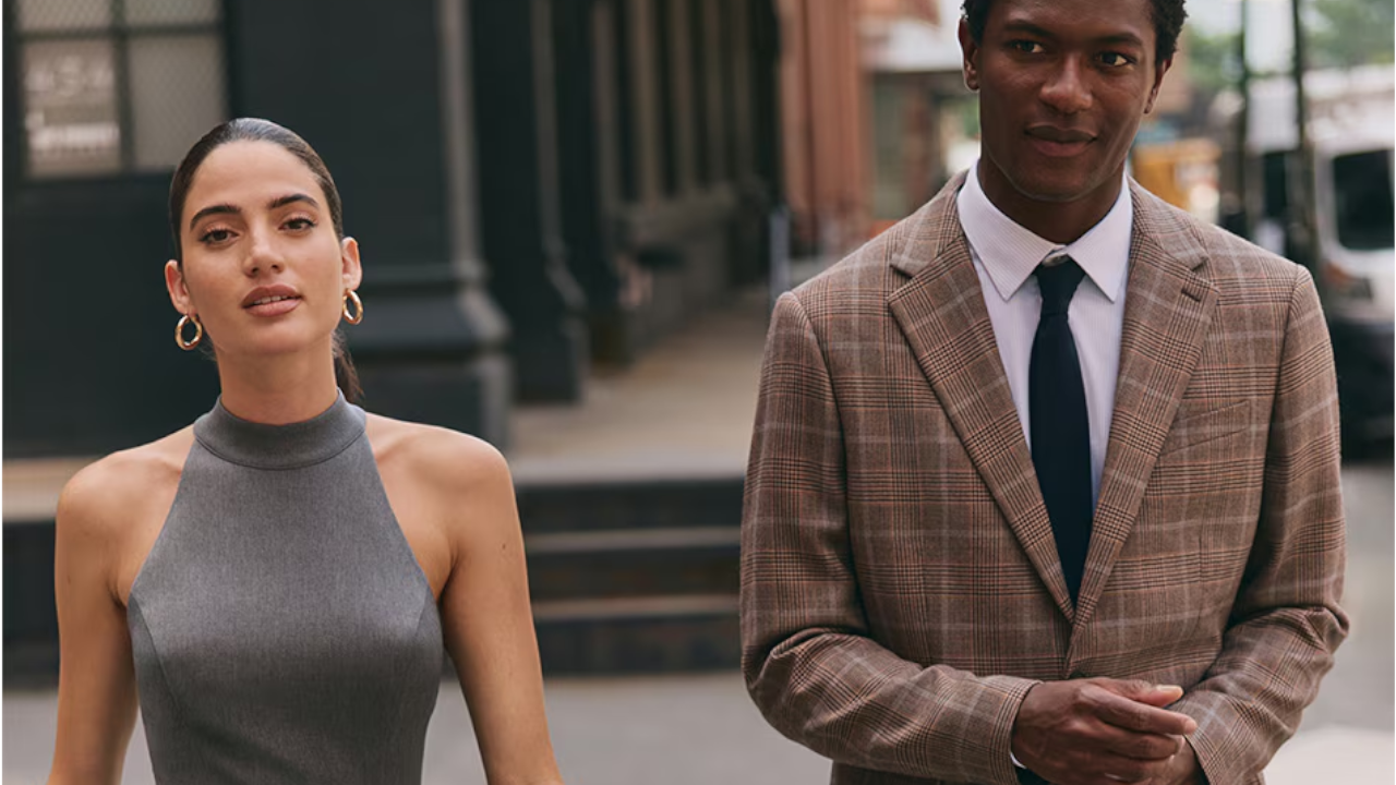 Woman in gray sleeveless top and man in plaid blazer with black tie standing on urban sidewalk