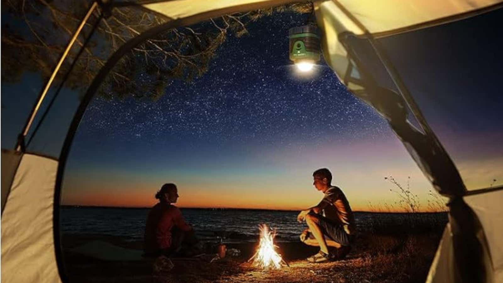 Two people sitting around a campfire on a beach at twilight with a starry sky overhead, viewed from inside a tent with a lantern hanging above