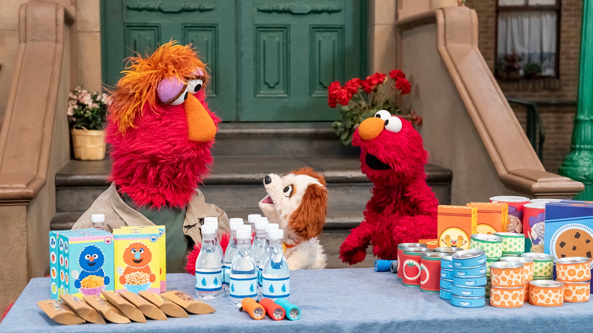 Elmo and other Muppets stand behind a table of emergency supplies including water bottles, canned goods, and flashlights on Sesame Street.
