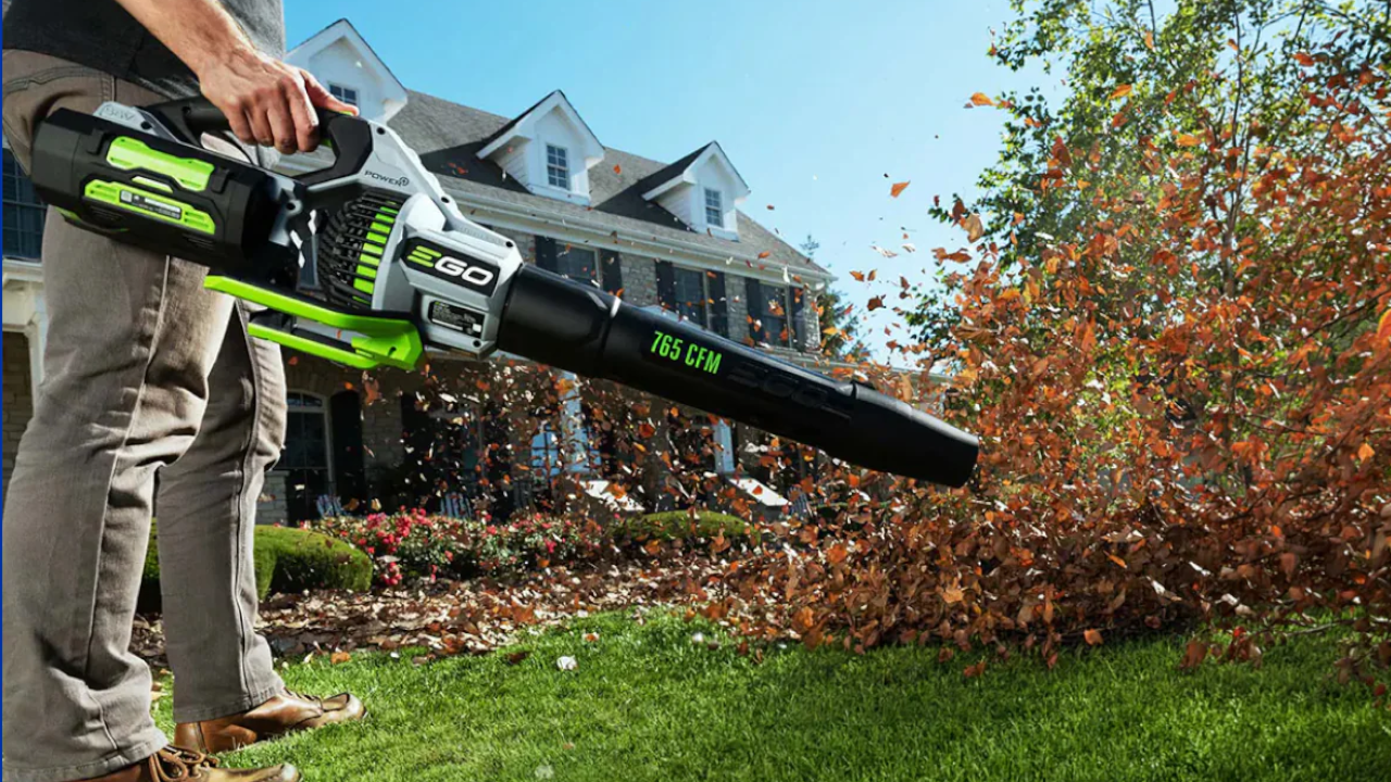 Person using an EGO 765 CFM leaf blower to clear autumn leaves from a lawn in front of a house