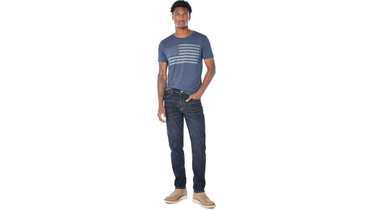 Man in navy American flag graphic t-shirt, dark jeans, and tan suede boots standing against white background