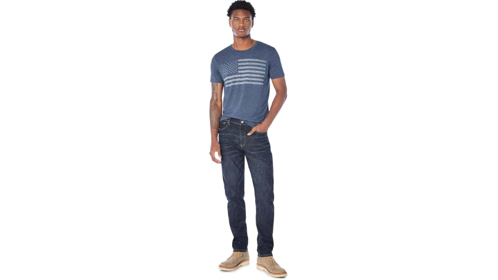 Man in navy American flag graphic t-shirt, dark jeans, and tan suede boots standing against white background