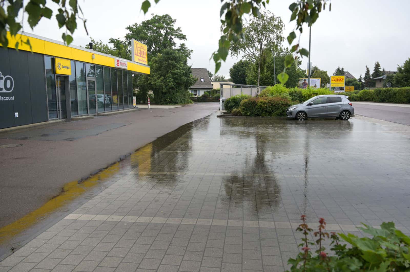 Wet pavement outside a Netto discount store with a gray car parked near green shrubs