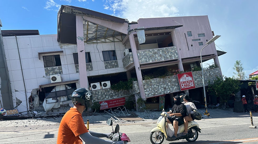 A scooter rides by a building partially collapsed in an earthquake