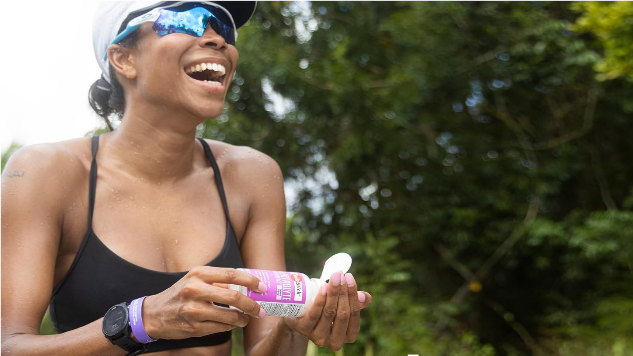 Smiling woman in sunglasses and cap holding a small bottle outdoors