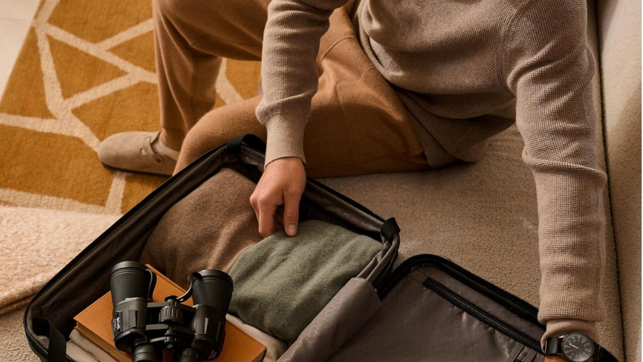 Person in beige sweater packing clothes and binoculars into an open black suitcase on a geometric patterned rug