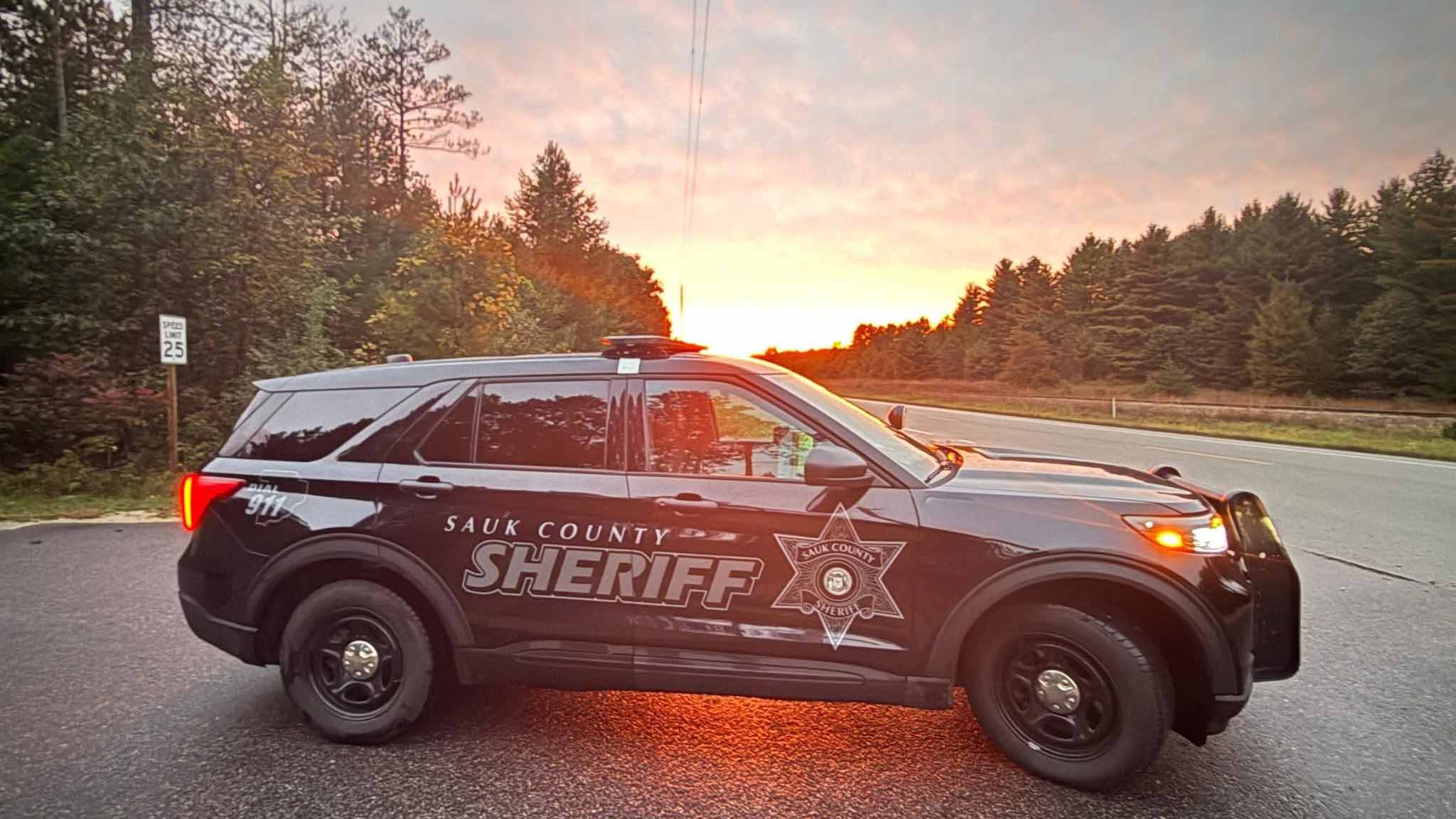 Black Sauk County Sheriff SUV parked on roadside at sunrise with tree-lined horizon