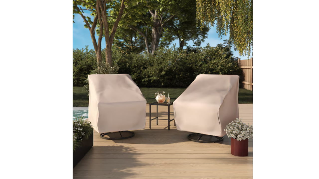 Two outdoor swivel chairs with beige protective covers flank a black side table with a pitcher on a wooden deck