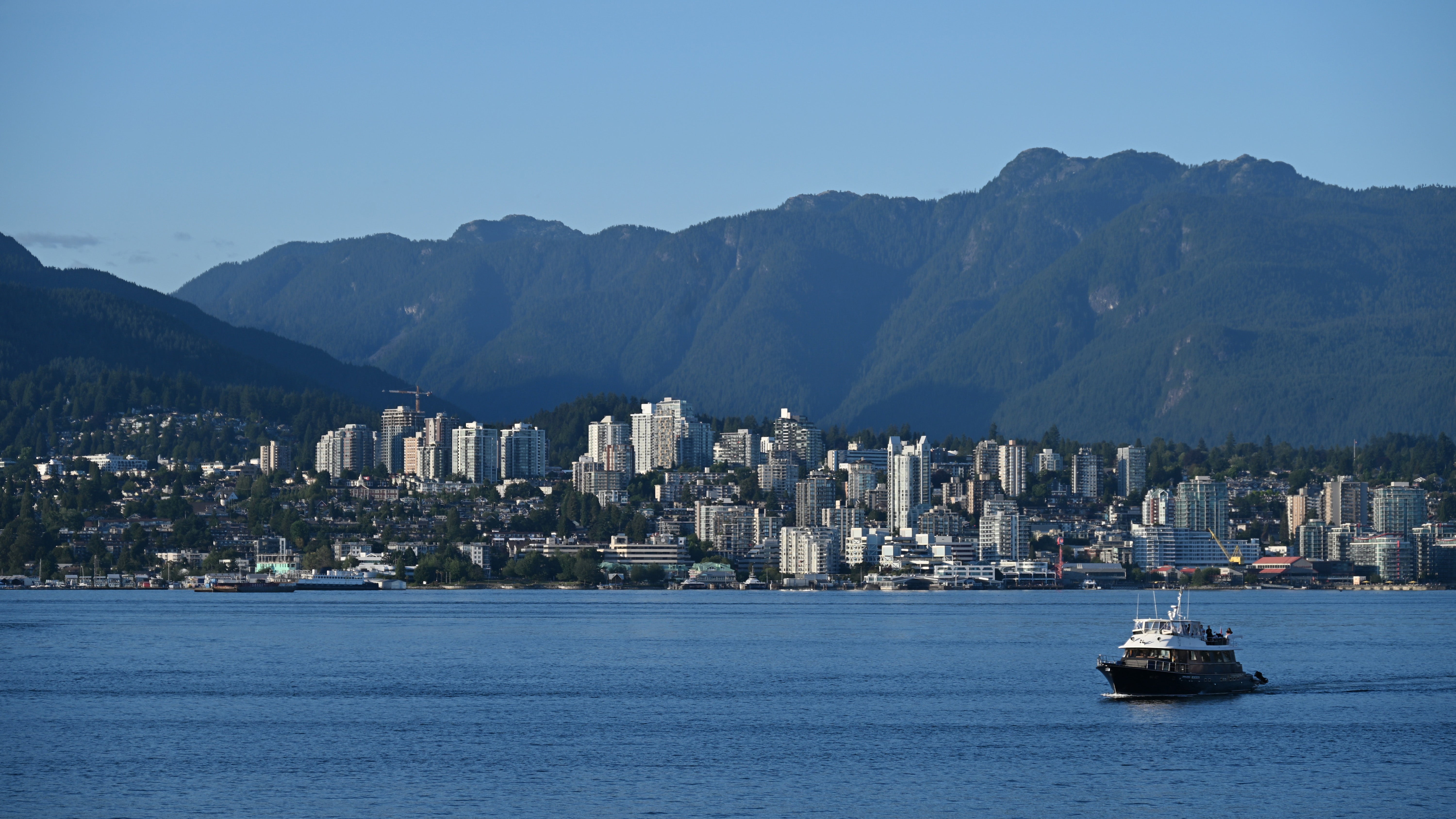 A ship sails on the water in front of the North Vancouver skyline. In the background, numerous skyscrapers and residential buildings in front of wooded mountains on a clear summer day. 
