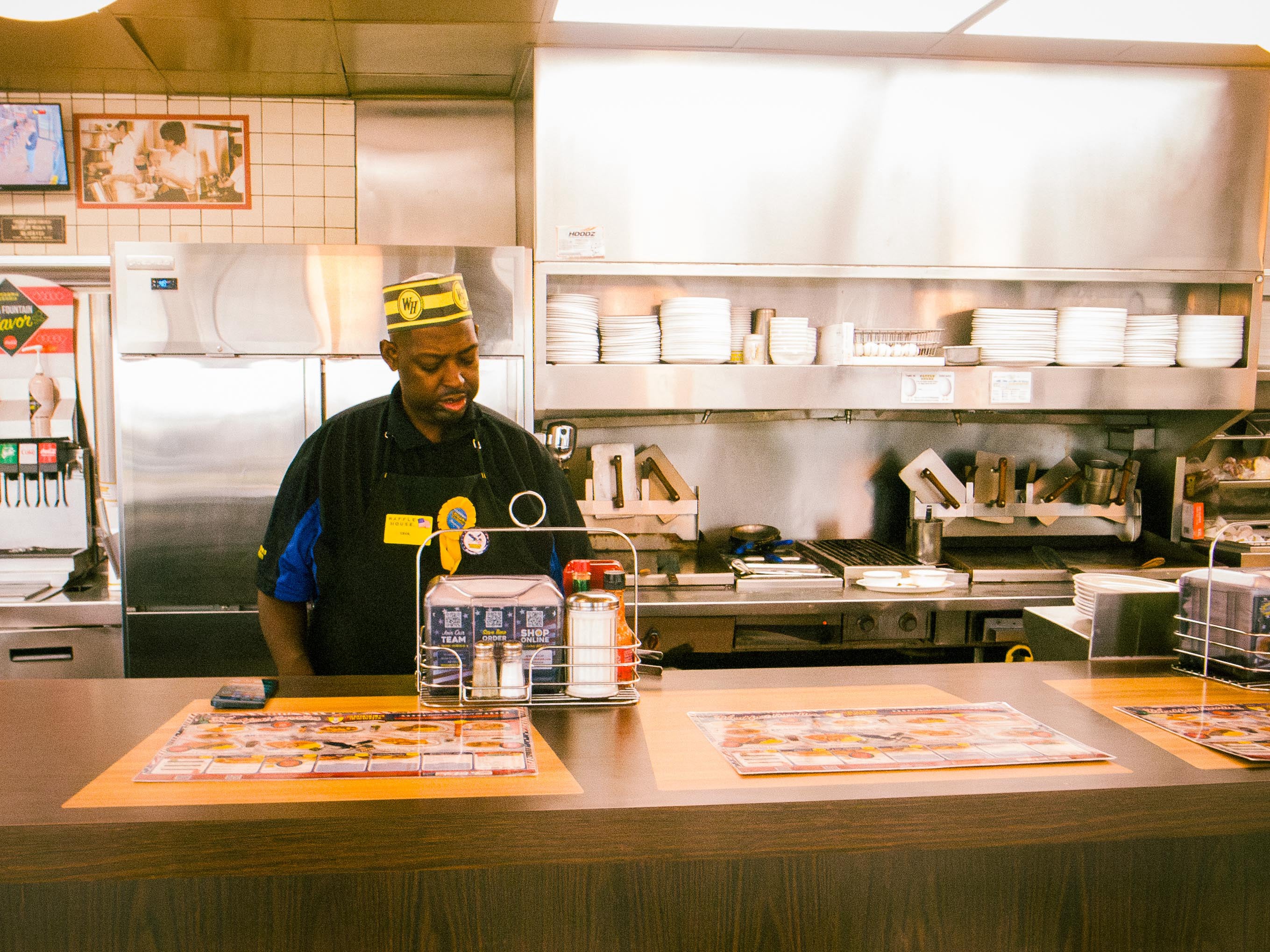 A Waffle House employee in a paper cap stands behind a counter with menus, in front of a grill and shelves of plates.