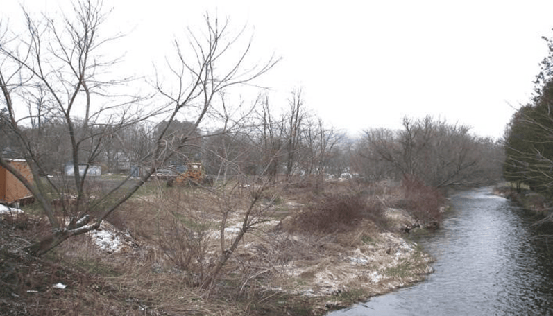Bare trees and brush line a river bank with patchy snow and a tractor visible in the distance