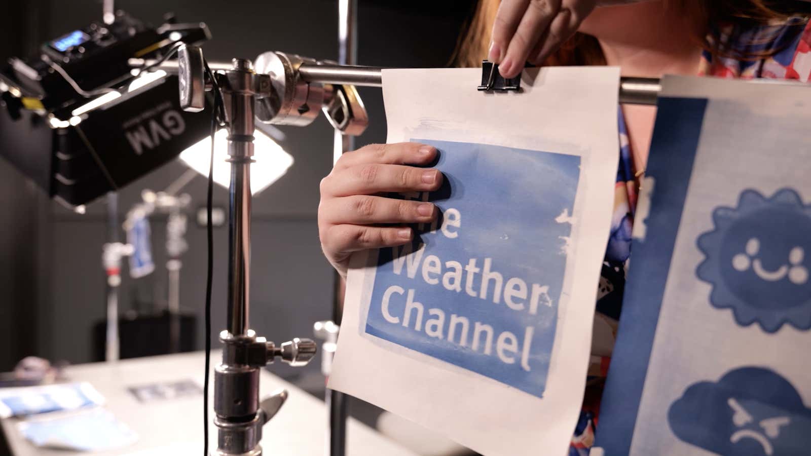 A hand clips a printed Weather Channel logo onto a photography stand next to studio lighting equipment