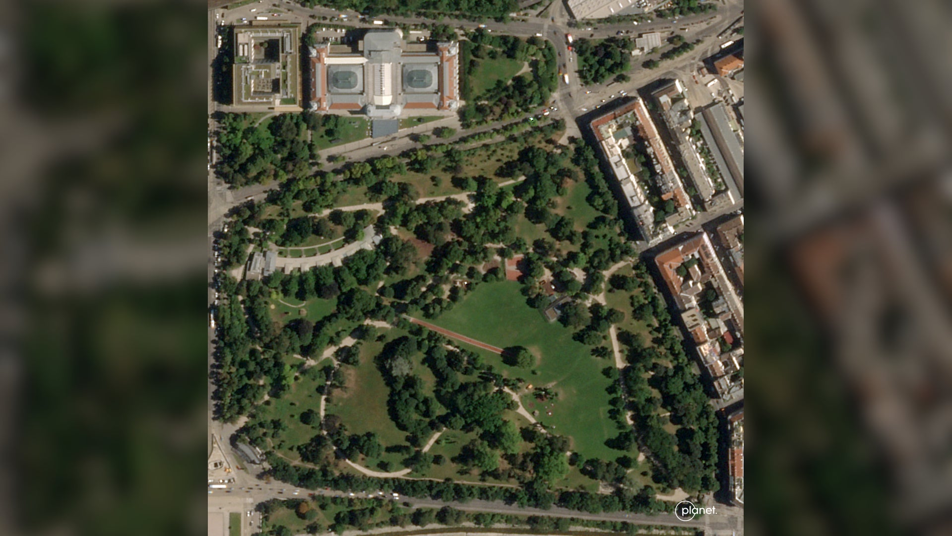 Satellite view of an urban park with winding paths and trees, adjacent to a large historic building and city blocks