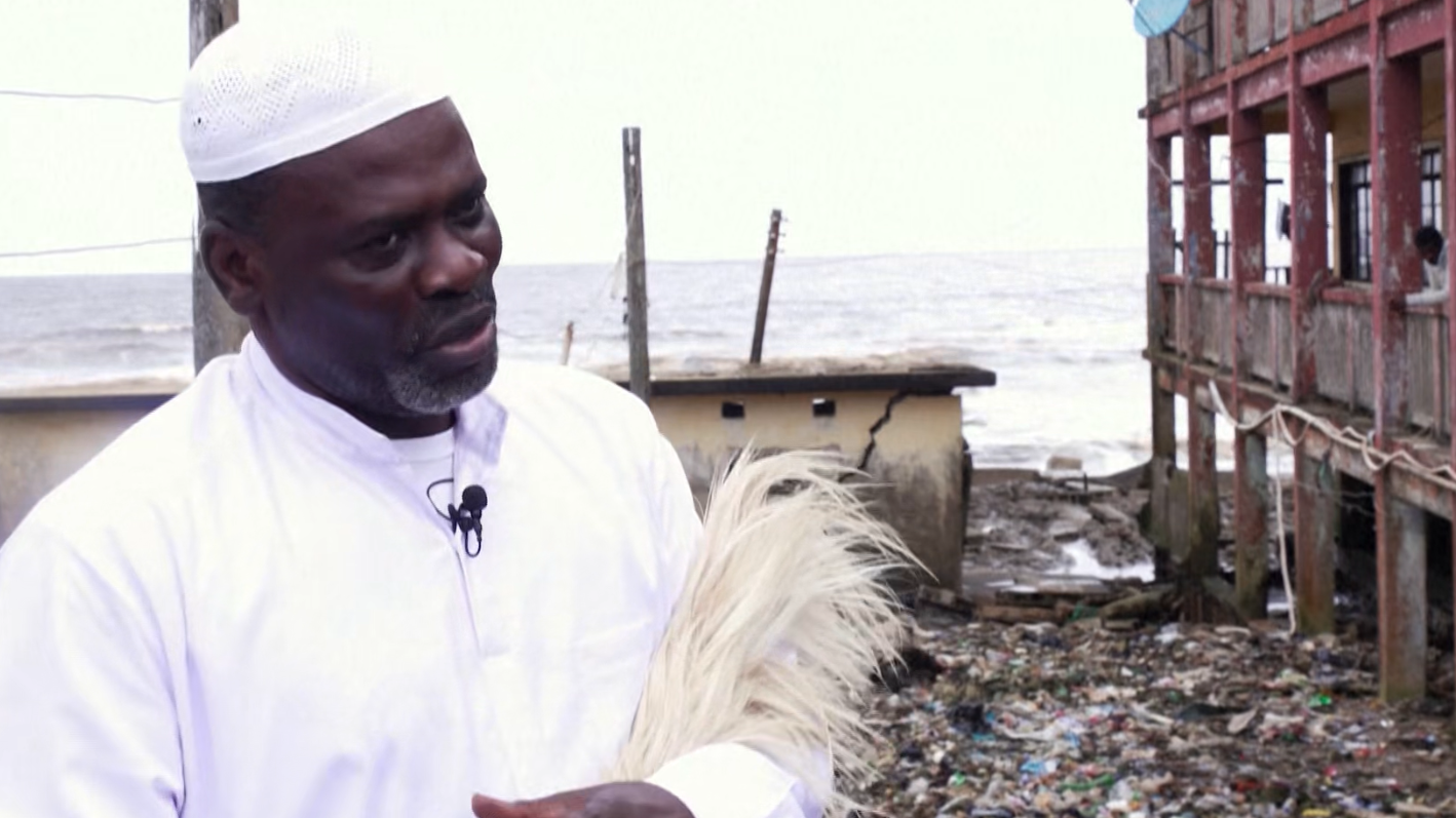 Man in white cap and traditional attire holding feathers near an eroding coastal building with debris and ocean waves behind him.