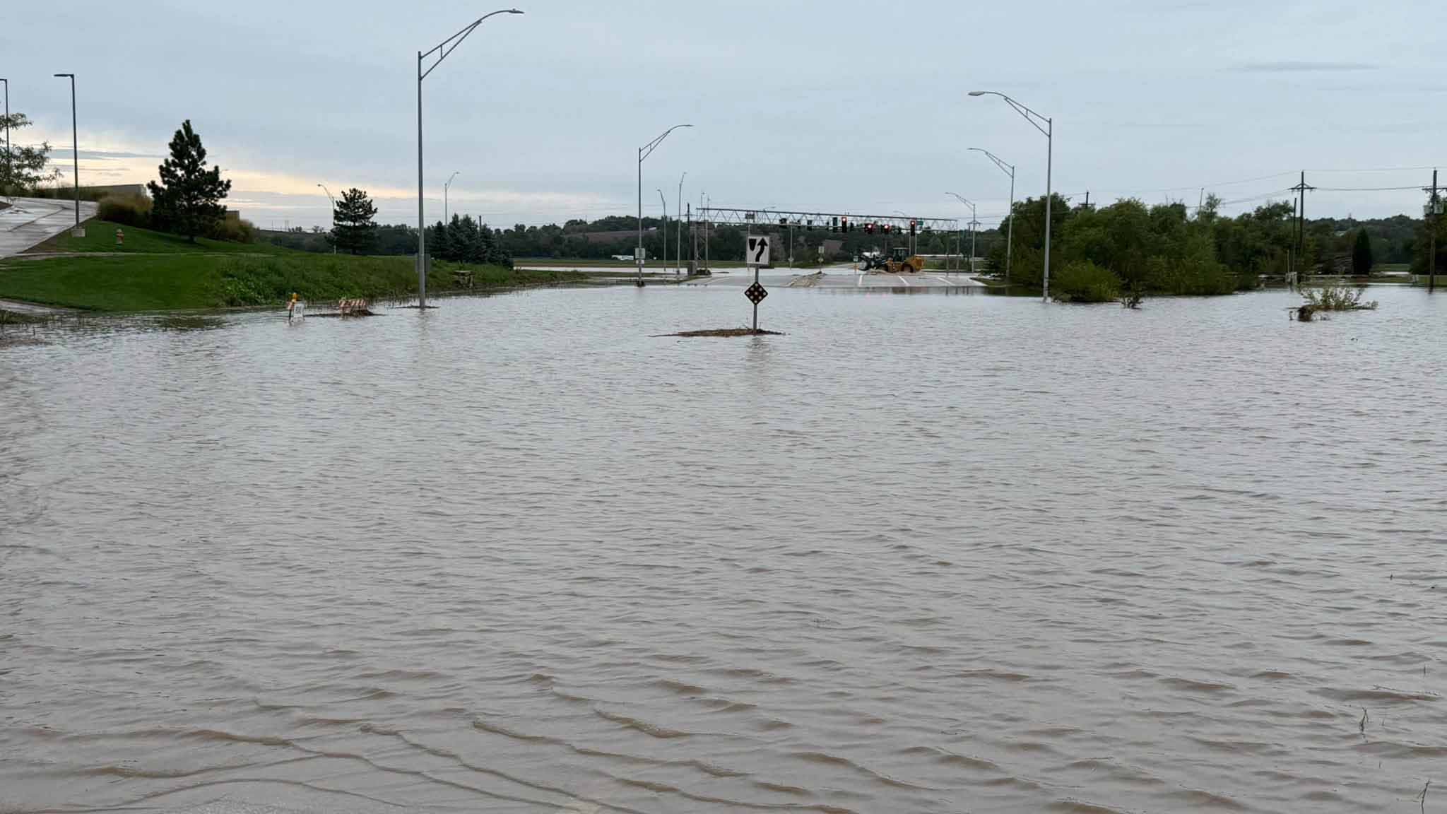 Flooded road intersection with submerged traffic signs, streetlights, and a tractor near distant traffic lights.