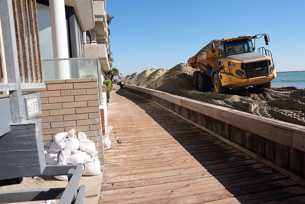 Yellow dump truck hauling sand along a wooden boardwalk beside beachfront buildings with sandbags stacked near the entrance