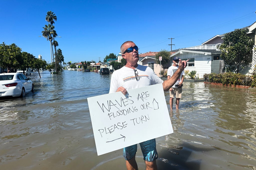 Man holding a sign reading "Waves are flooding our street, please turn" while standing in a flooded residential street with submerged cars and palm trees