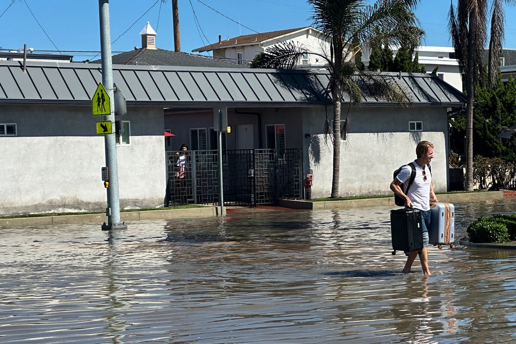 Man wades barefoot through flooded street carrying two suitcases near a gray house with palm trees