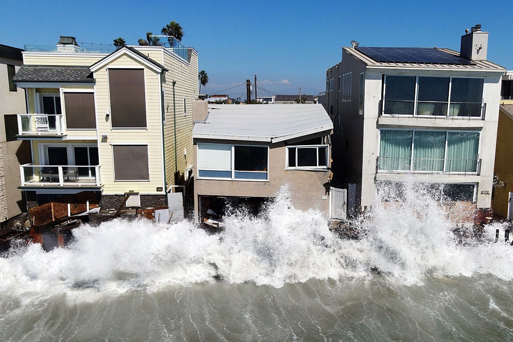 Large ocean waves crash against beachfront houses, splashing water onto the buildings' lower floors