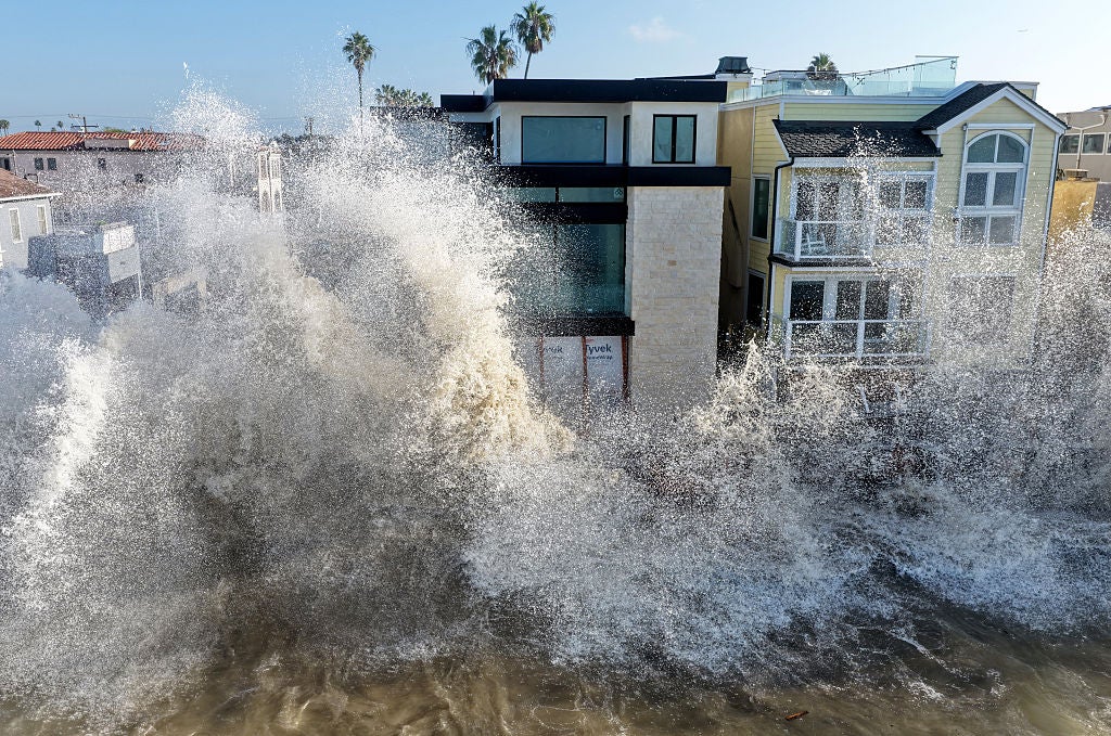 Large ocean wave crashes against beachfront houses, spraying high above the rooftops