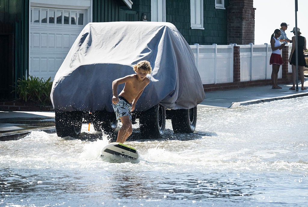 Shirtless boy surfing on a flooded residential street past a covered truck and onlookers
