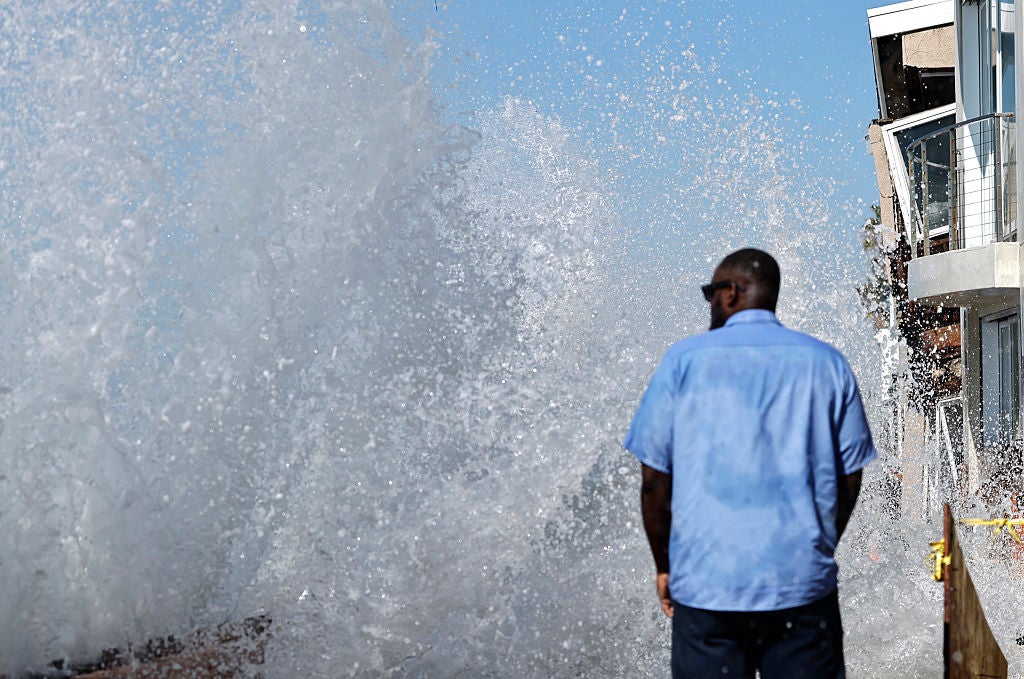 Man in blue shirt watches large ocean wave crash against seawall near coastal buildings