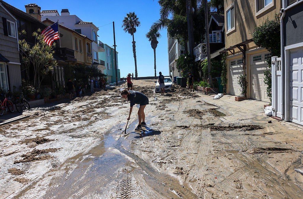 People shoveling mud from a flooded residential street lined with houses and palm trees after a storm