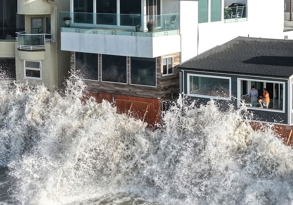 Large ocean wave crashes against oceanfront homes as two people watch from a window above