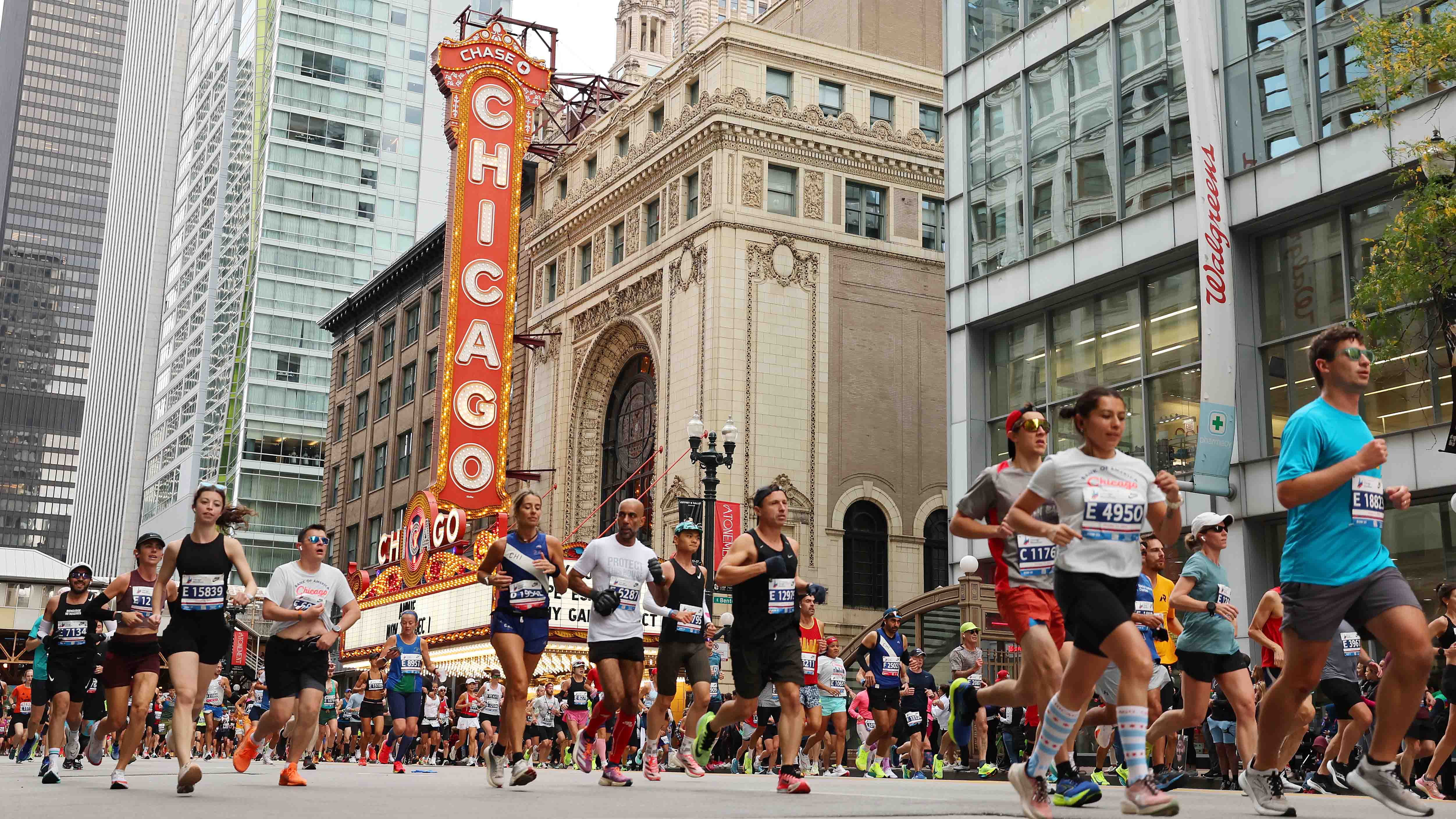 Marathon runners with race bibs pass the Chicago Theatre marquee sign amid downtown skyscrapers