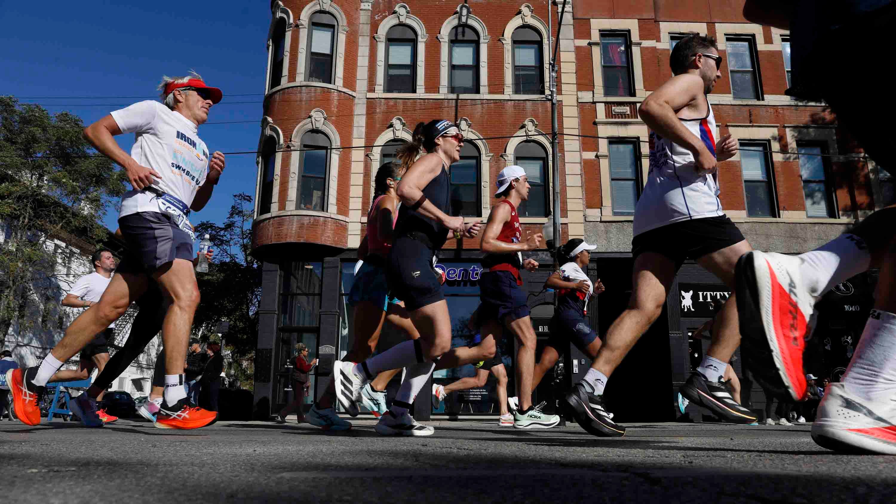 Marathon runners race down a city street past brick buildings with arched windows under a clear blue sky