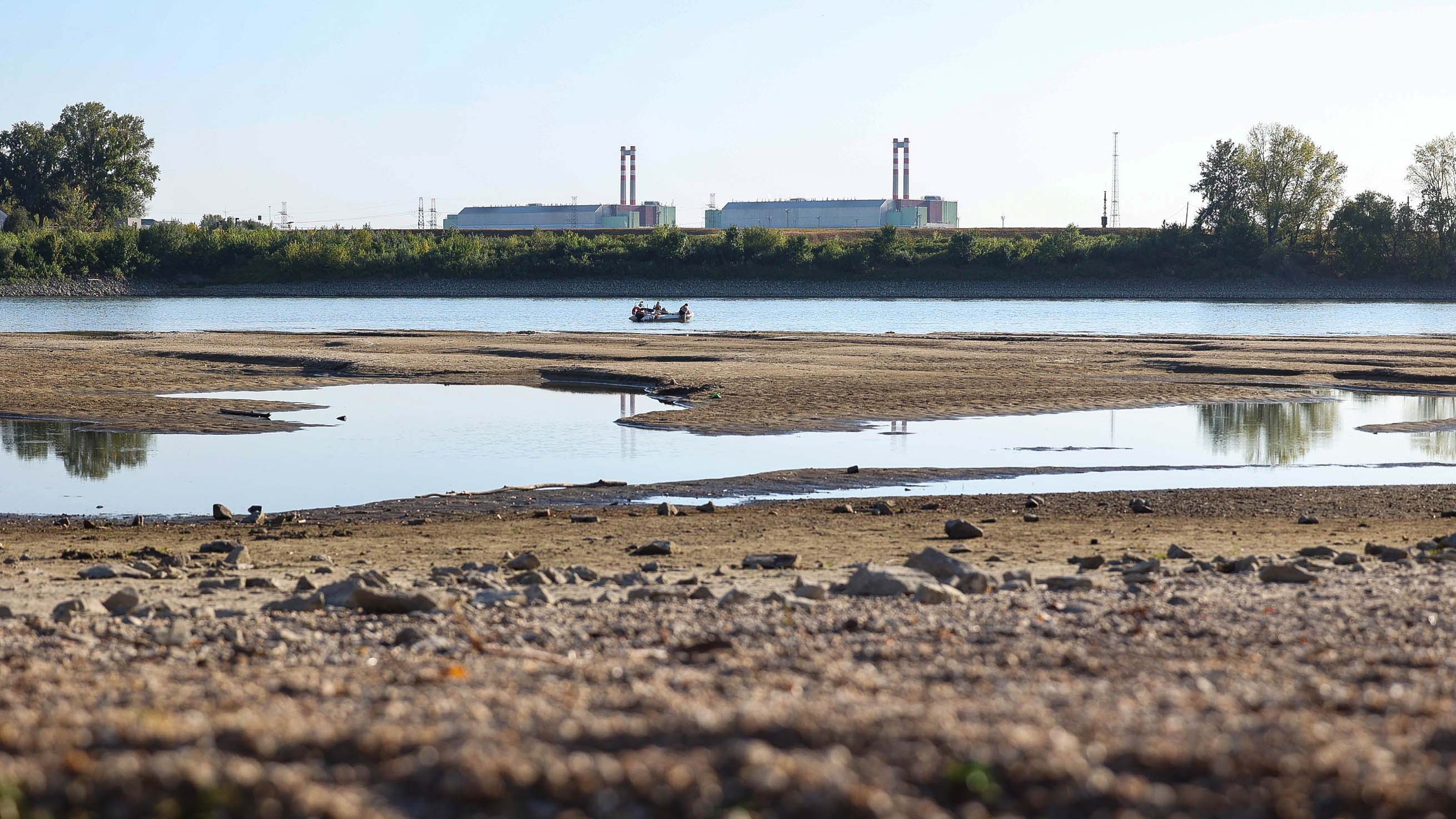 Exposed sandbars and low water levels on a river, with a small boat and industrial plant with twin smokestacks in the background