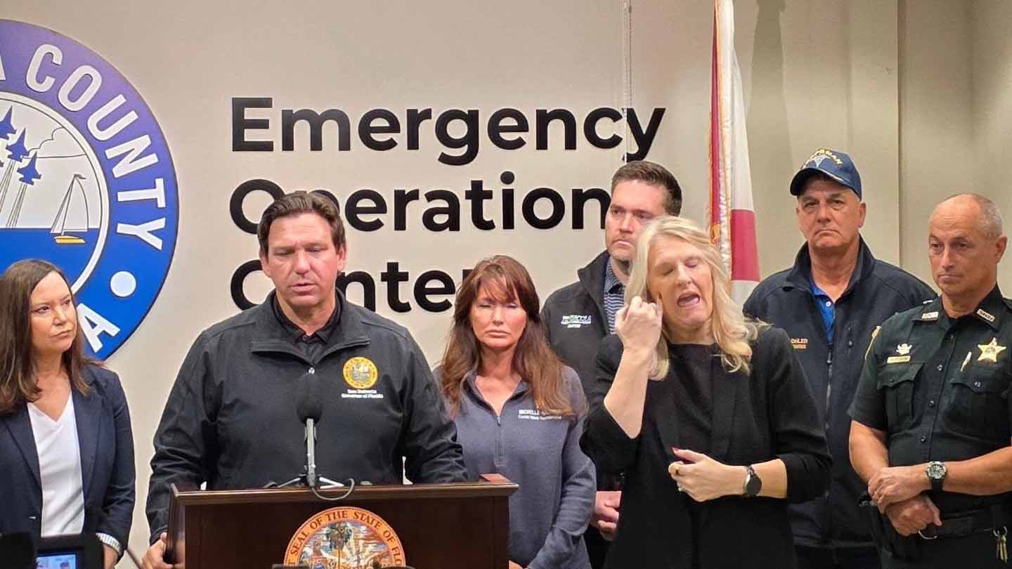 Man speaking at podium during press conference at county Emergency Operations Center, surrounded by officials and a sign language interpreter.