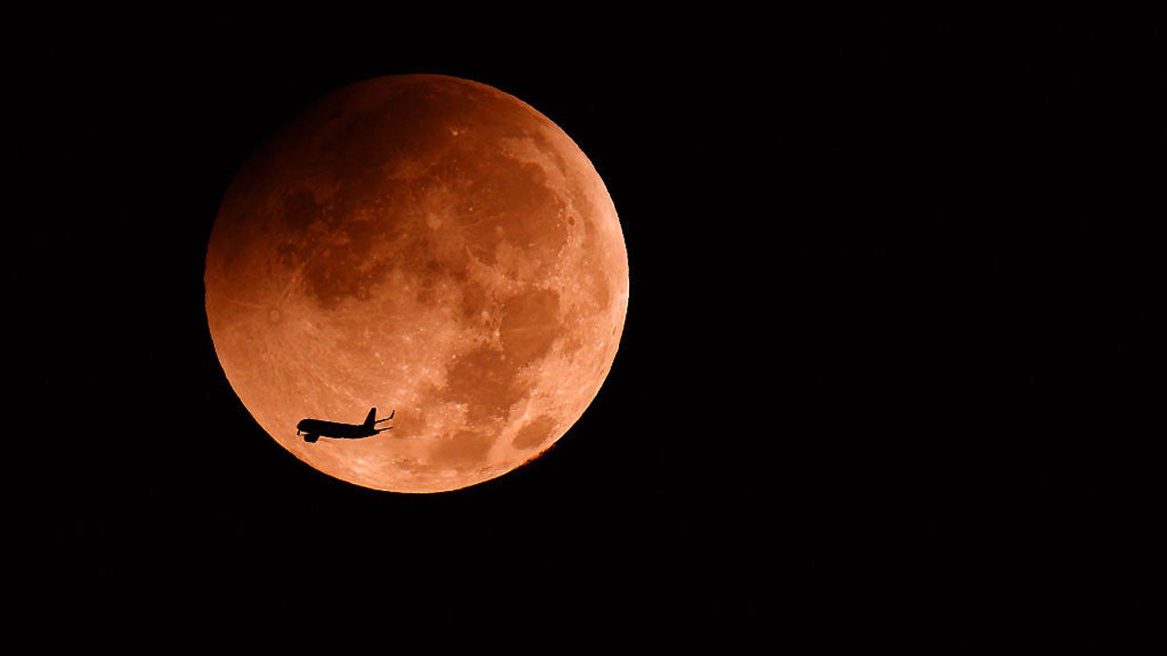 Silhouette of an airplane flying across a glowing orange full moon against a black night sky