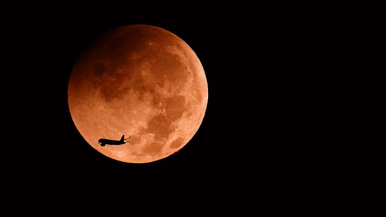 Silhouette of an airplane flying across a glowing orange full moon against a black night sky
