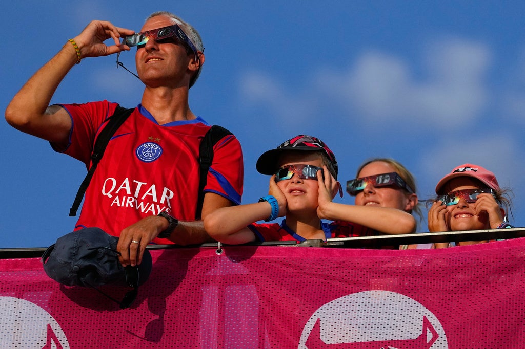 A man wearing a Paris Saint-Germain jersey and three children wearing solar eclipse glasses look up at the sky from behind a pink barrier