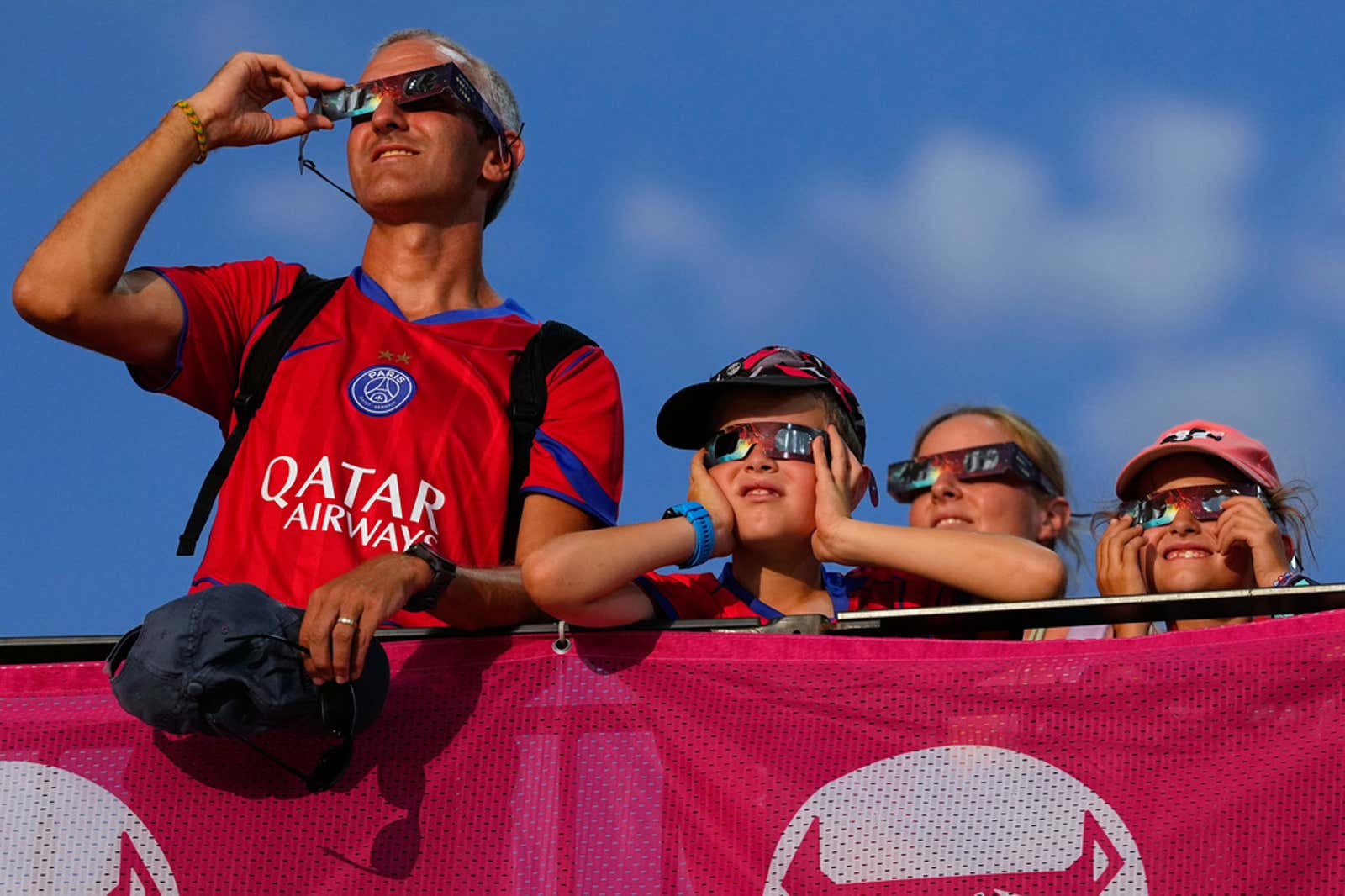 A man wearing a Paris Saint-Germain jersey and three children wearing solar eclipse glasses look up at the sky from behind a pink barrier