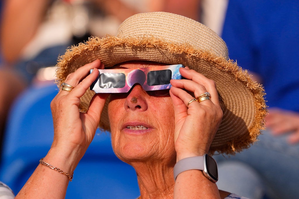 Woman in a straw hat holding solar eclipse viewing glasses up to her eyes outdoors.