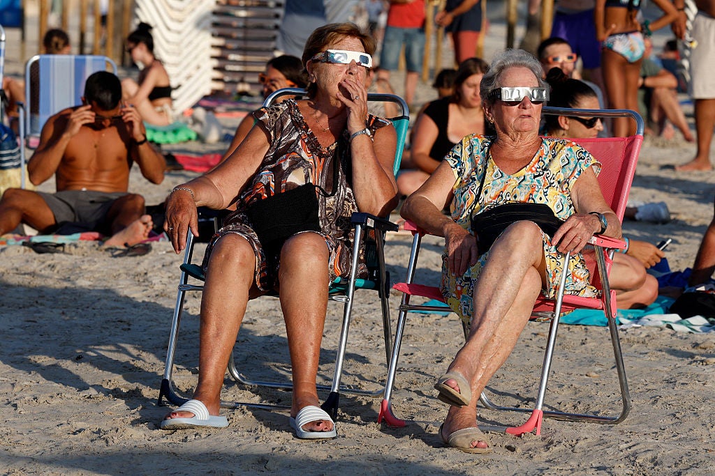 Two women wearing solar eclipse glasses sit in beach chairs on a sandy beach with other sunbathers